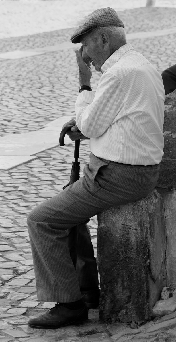 An elderly gentleman sits with his umbrella on a wall at the side of a cobblestone street.