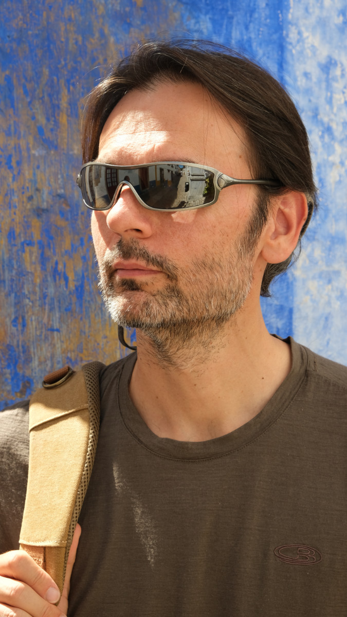 A portrait of a man wearing sunglasses, standing against a weathered blue wall